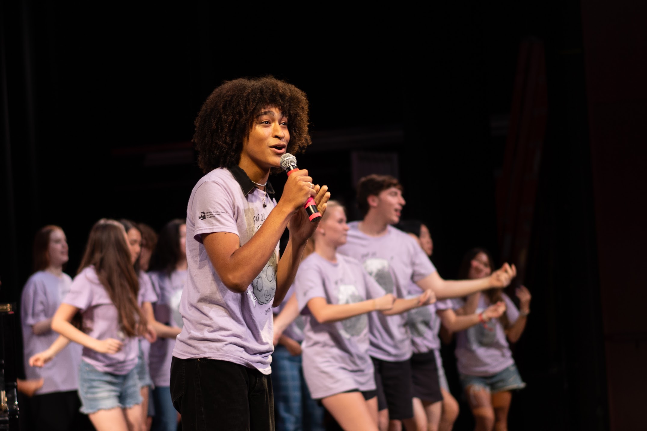 A moment from the FAP 28 pageant. A dark-skinned person with curly hair wearing a purple FAP shirt sings into a microphone while other FAP students dance in the background.